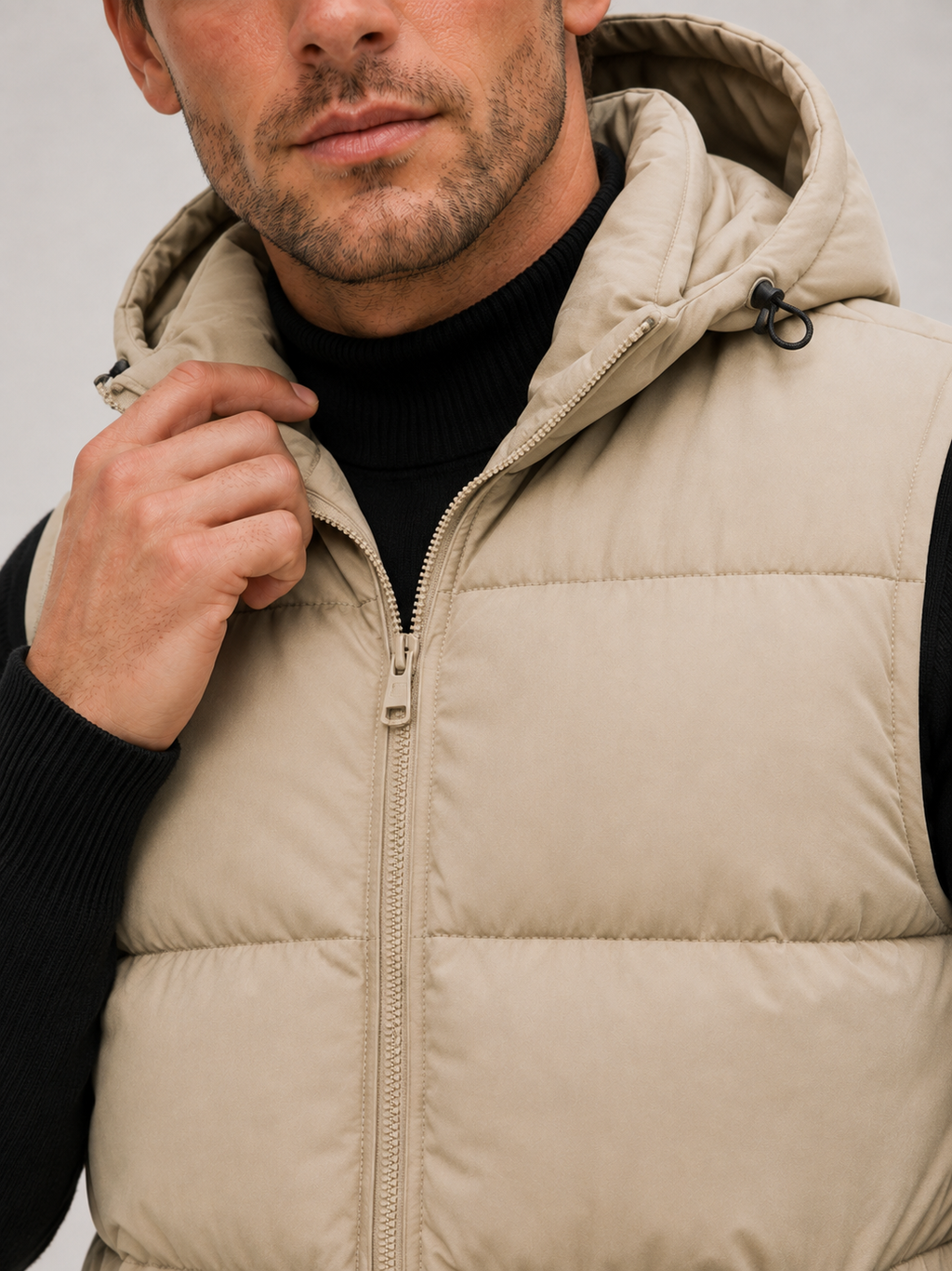 Close-up of a beige hooded puffer vest showing the thick quilted insulation, full zipper, hood, pockets, armholes, and stitching