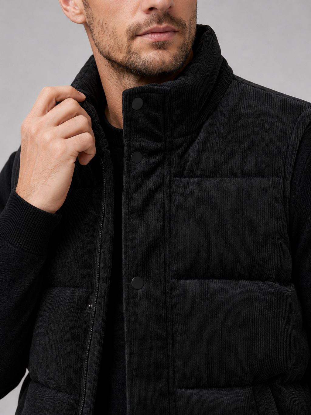 Close-up of a black corduroy puffer vest showing the ribbed fabric texture, thick padding, stand collar, zipper, pockets, armholes, and stitching