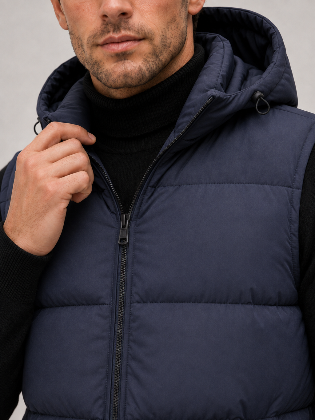 Close-up of a navy hooded puffer vest showing the thick quilted insulation, full zipper, hood, pockets, armholes, and stitching