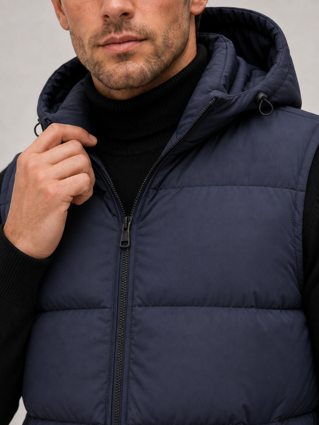 Close-up of a navy hooded puffer vest showing the thick quilted insulation, full zipper, hood, pockets, armholes, and stitching
