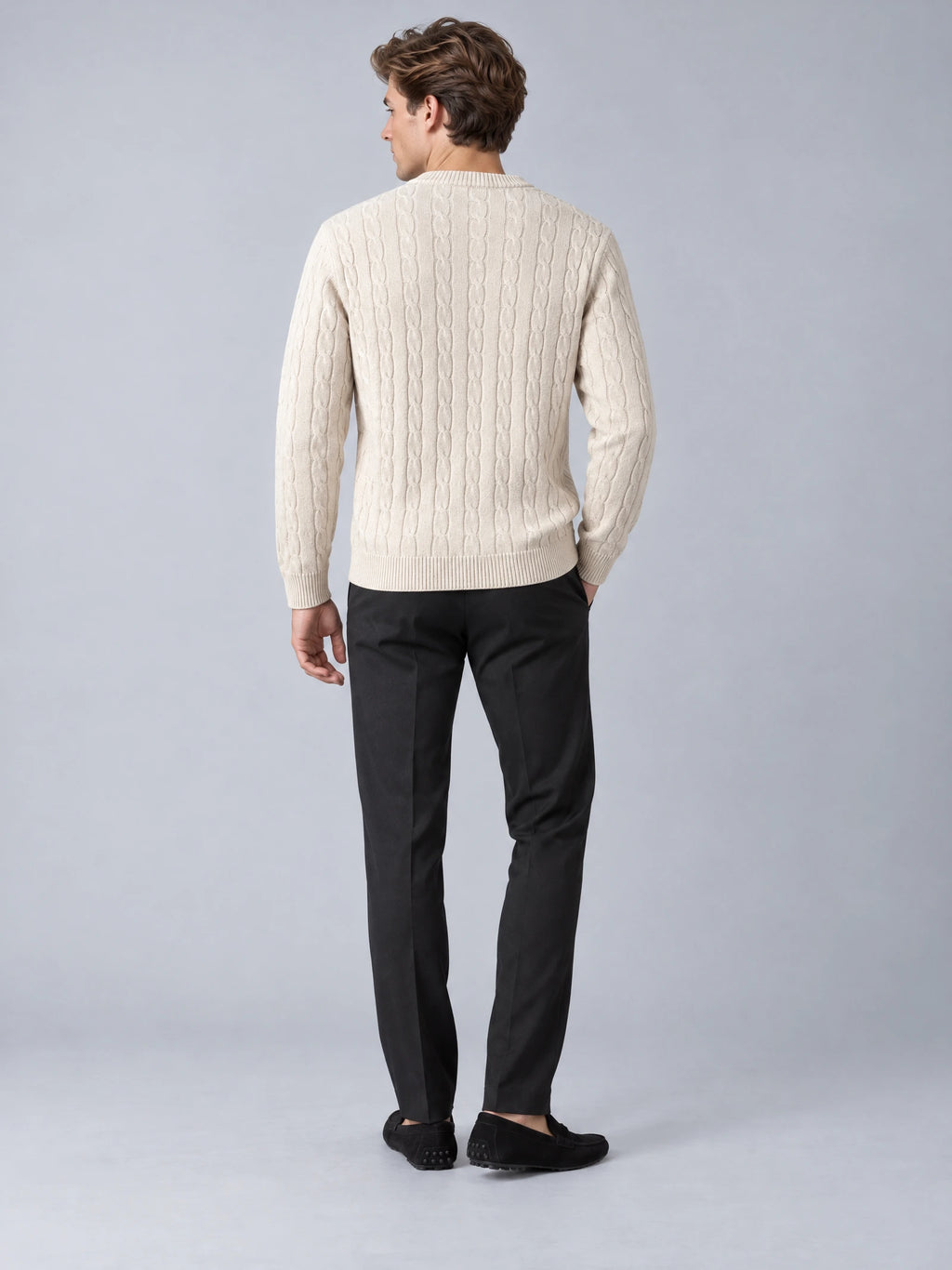 Back view of a model wearing a Beige cable knit sweater with long sleeves and ribbed crewneck, cuffs, and hem.