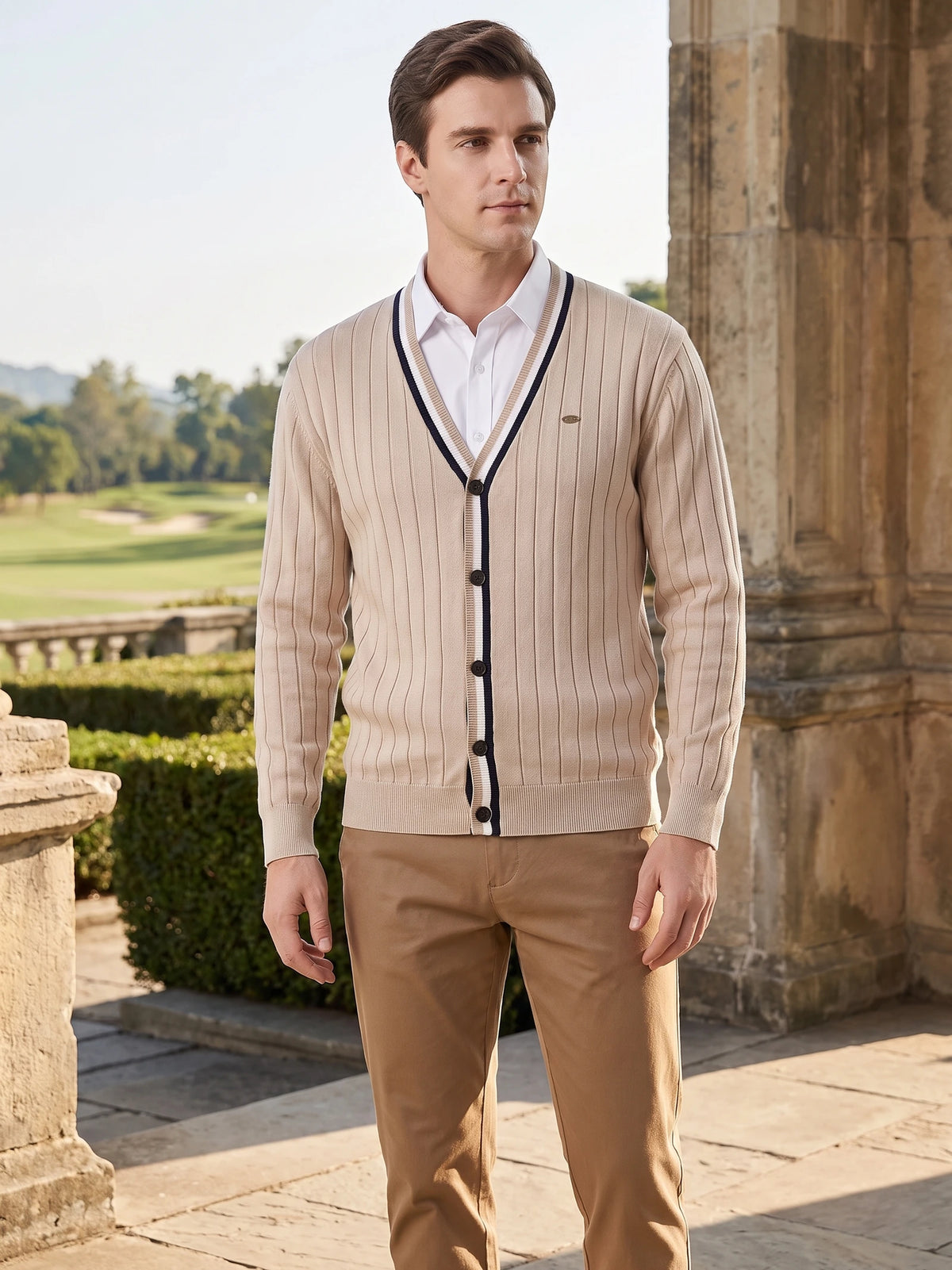 Model wearing a Beige cotton Cardigan Sweater with a ribbed V-neck and button front beside a stone terrace overlooking a landscaped lawn.