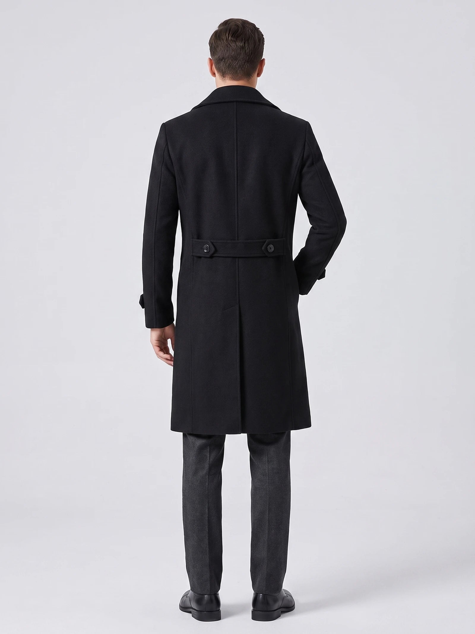Rear studio view of a model wearing a Black knee-length trench coat with a buttoned back belt and center vent.