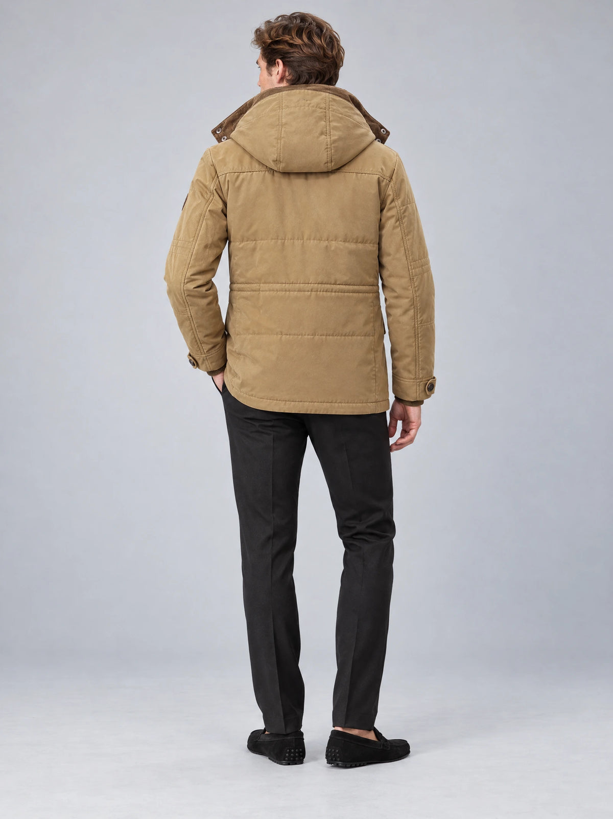 Rear studio view of a model wearing a Khaki heavy winter jacket with a hood, quilted back panels, and buttoned cuffs.