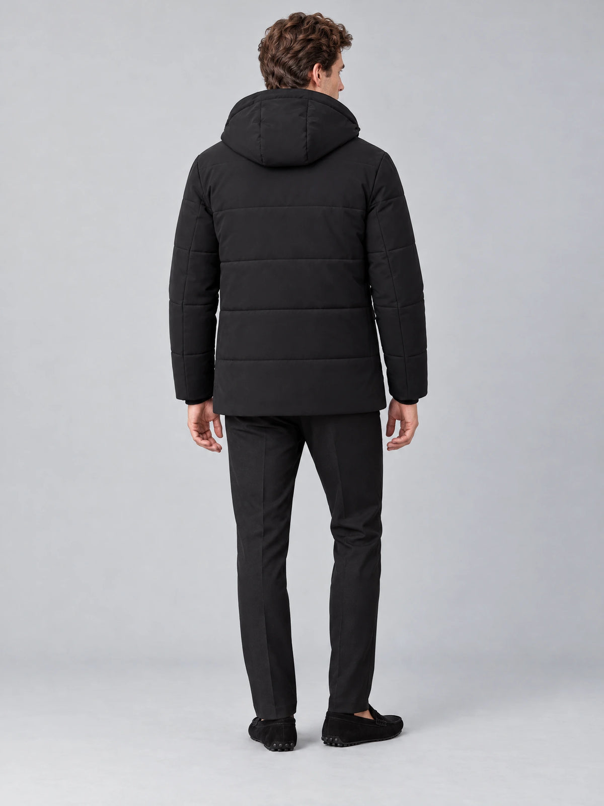 Rear studio view of a model wearing a Black Heavy Winter Jacket with a padded hood and horizontal quilted back panels.