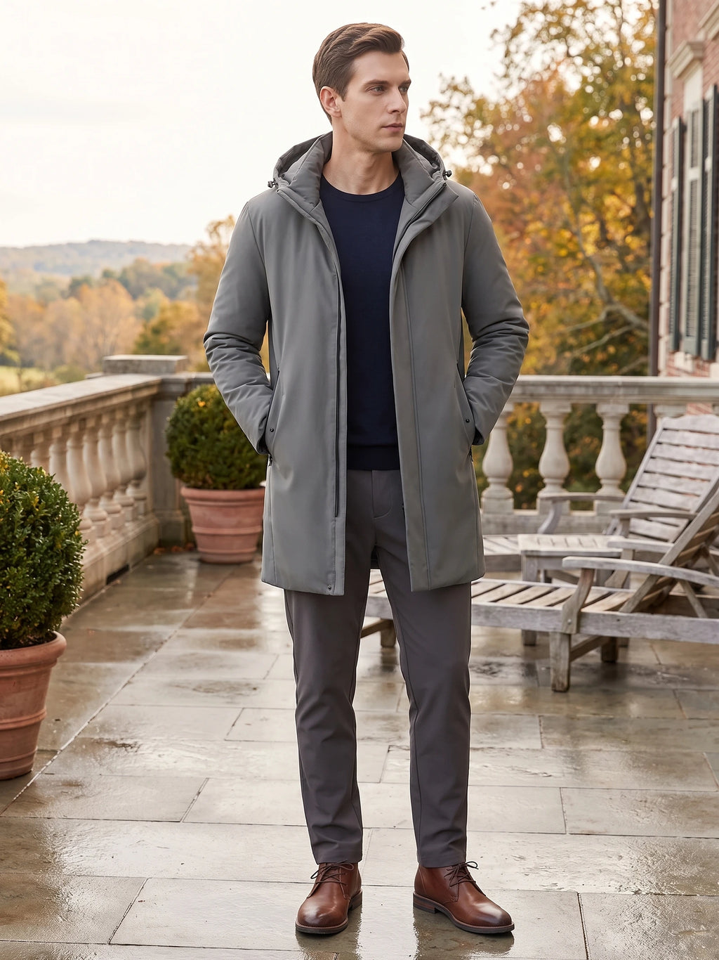 Model wearing a Gray hooded trench coat with hands in pockets on a terrace beside autumn trees.
