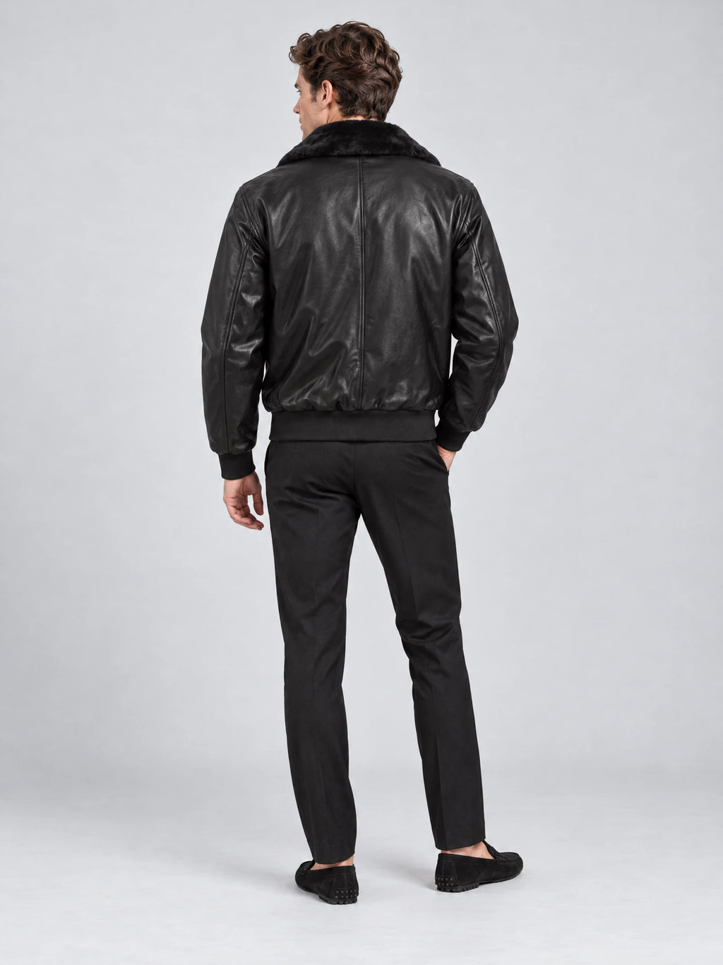 Rear studio view of a model wearing a Black leather bomber jacket with a plush collar, back seam, and ribbed hem.
