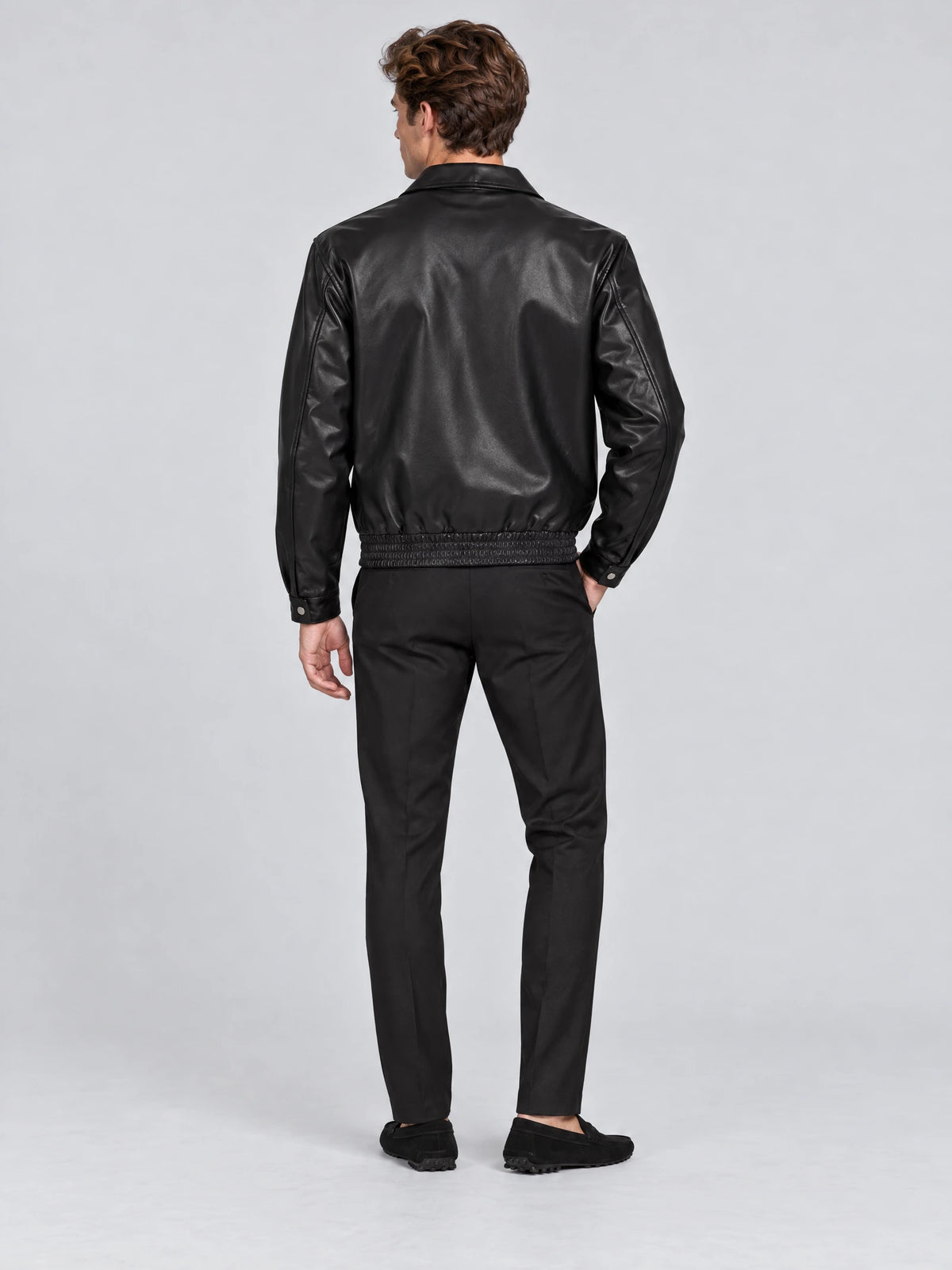 Rear studio view of a model wearing a Black leather bomber jacket with a lapel collar, elasticated hem, and buttoned cuffs.