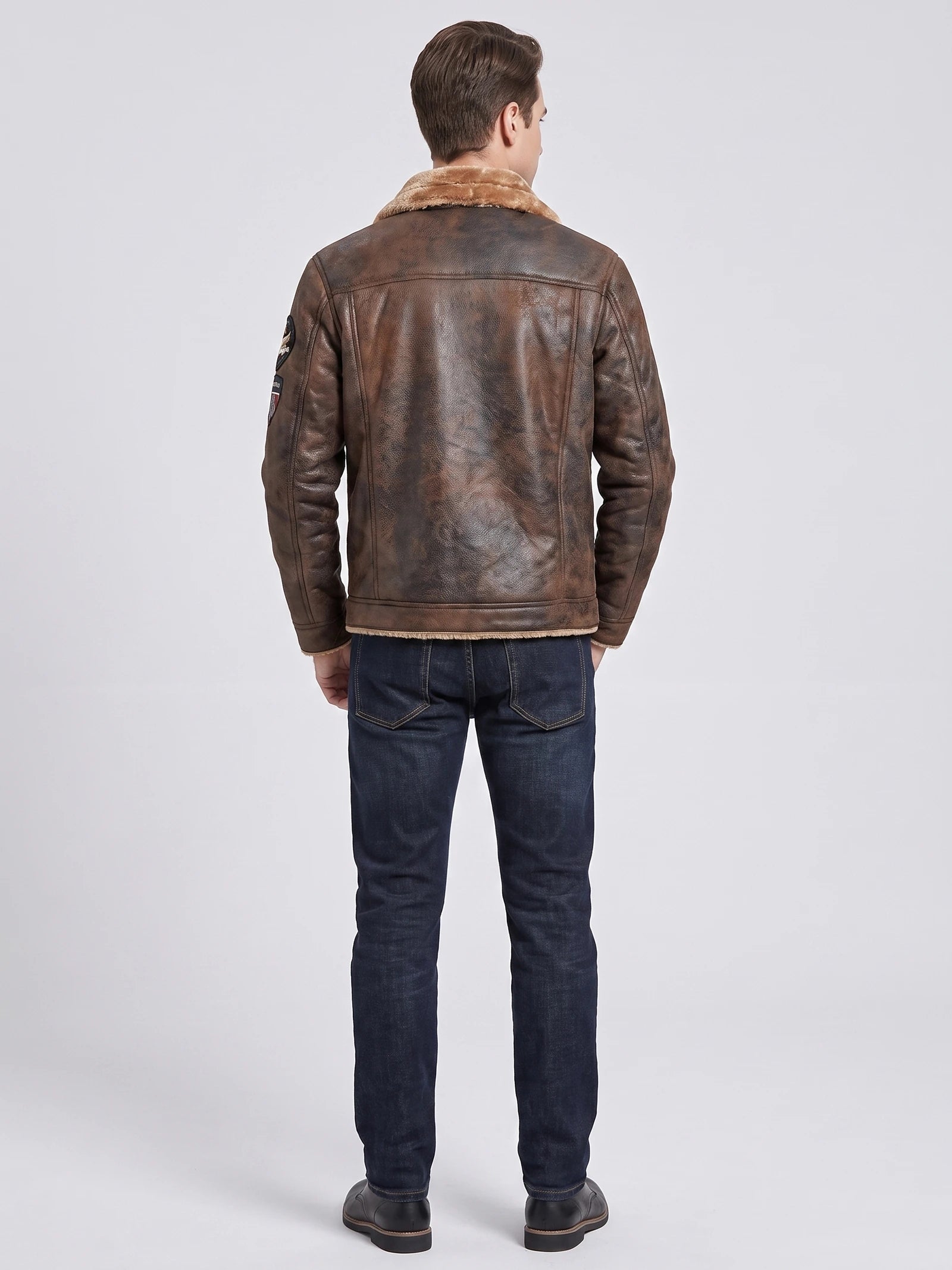 Rear studio view of a model wearing a Brown leather jacket with a fleece collar, paneled back, and sleeve patch.