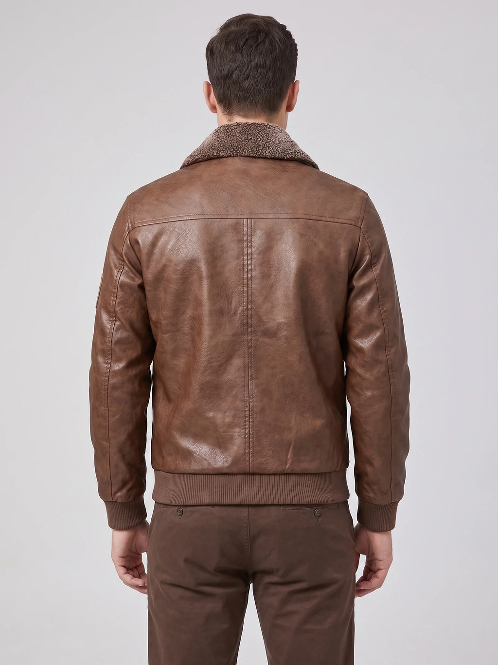 Rear view of a model wearing a Brown leather jacket with a textured fur collar, back seam detailing, and ribbed hem and cuffs.