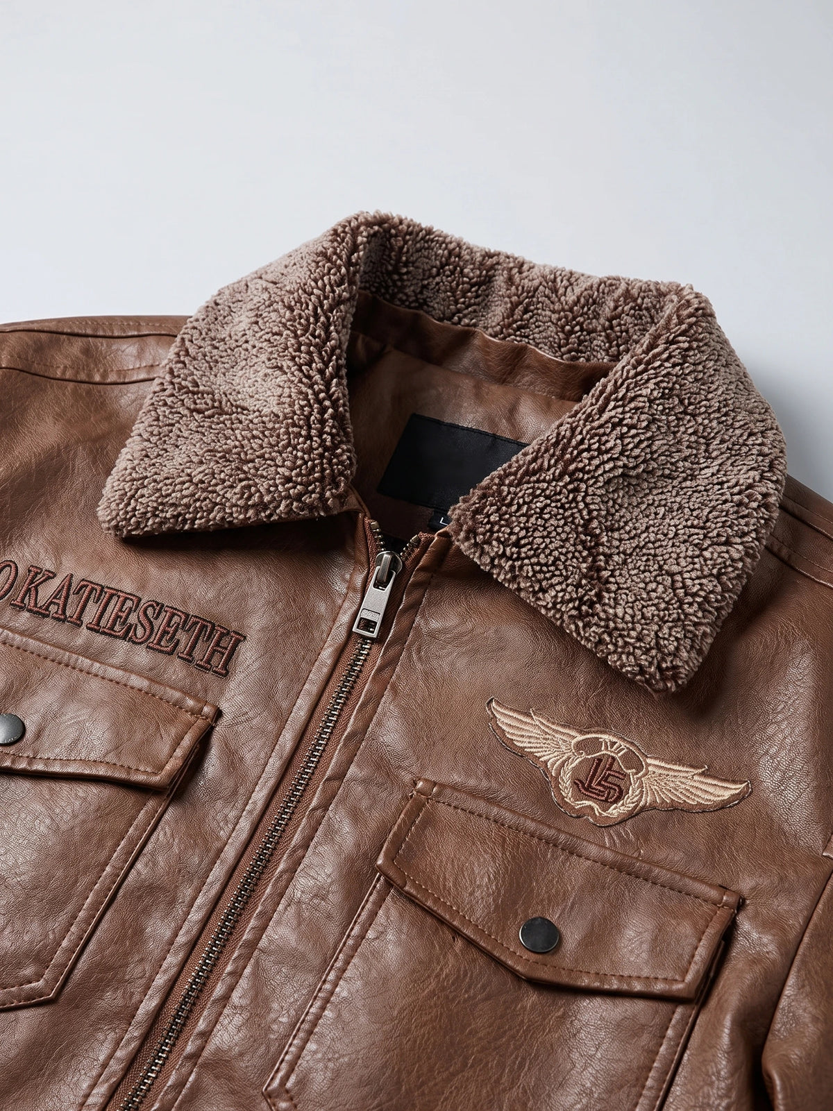 Close-up of a Brown Leather Jacket with a textured fur collar, front zipper, embroidered chest patches, and snap-flap pockets.