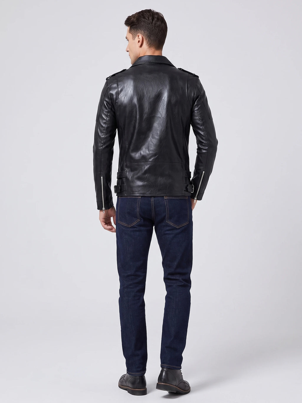 Rear studio view of a model wearing a Black PU leather motorcycle jacket with shoulder tabs, back seams, adjustable waist tabs, and zippered cuffs.