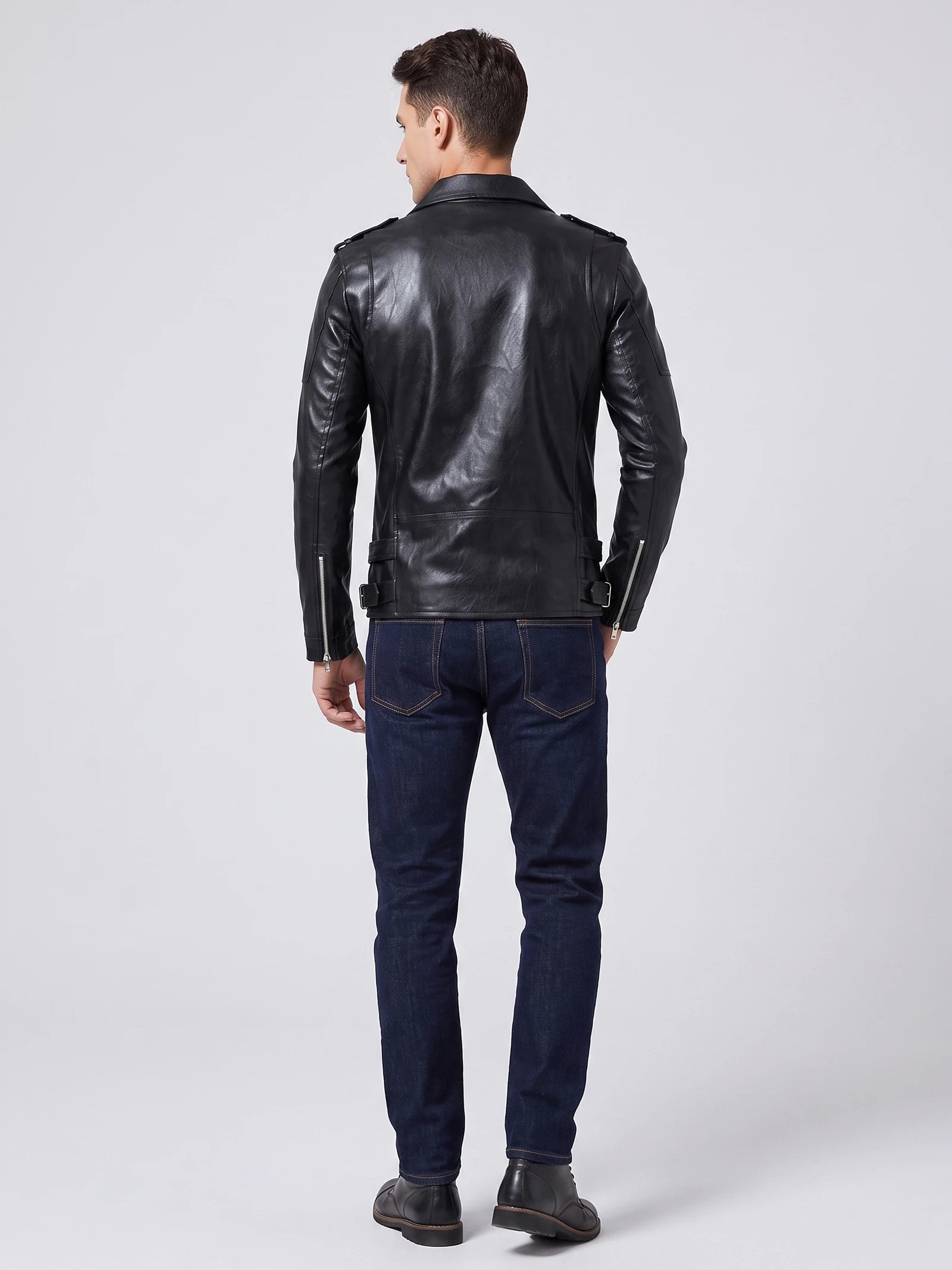 Rear studio view of a model wearing a Black PU leather motorcycle jacket with shoulder tabs, back seams, adjustable waist tabs, and zippered cuffs.