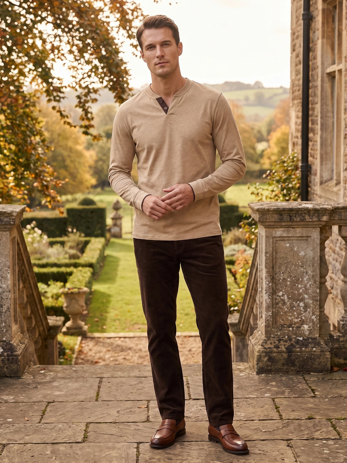 Model wearing a Khaki long sleeve Henley shirt on a stone terrace overlooking a landscaped garden.