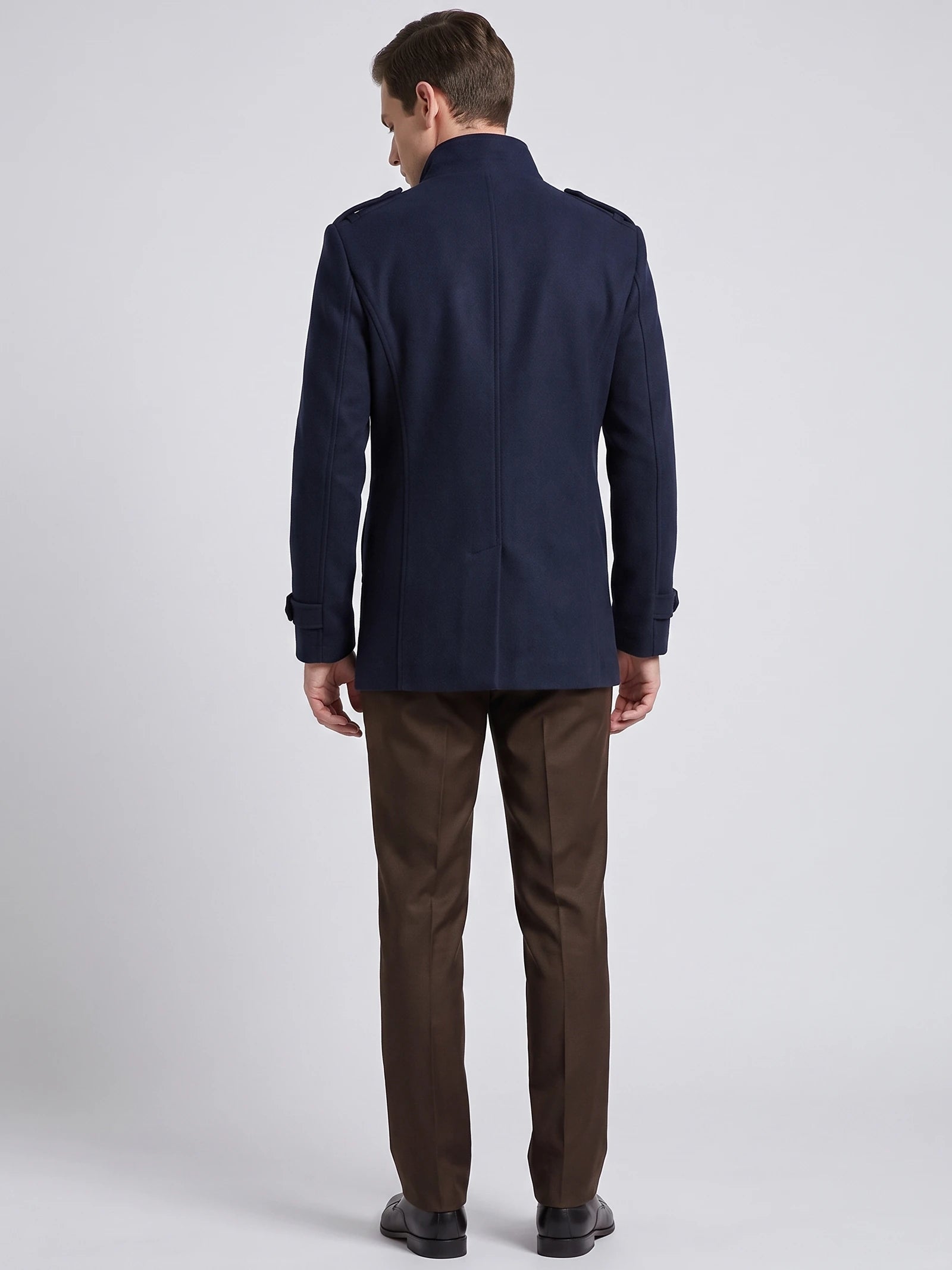 Rear studio view of a model wearing a Navy wool overcoat with a stand collar, shoulder tabs, cuff straps, and a center back seam.