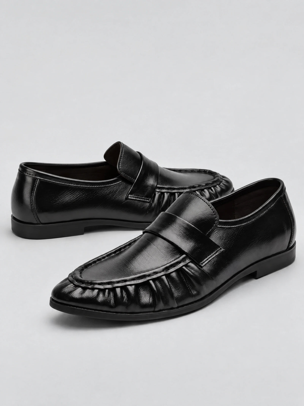 Black ruched leather loafers shown as a pair in a studio product photo, featuring slip-on straps and gathered toe detailing.