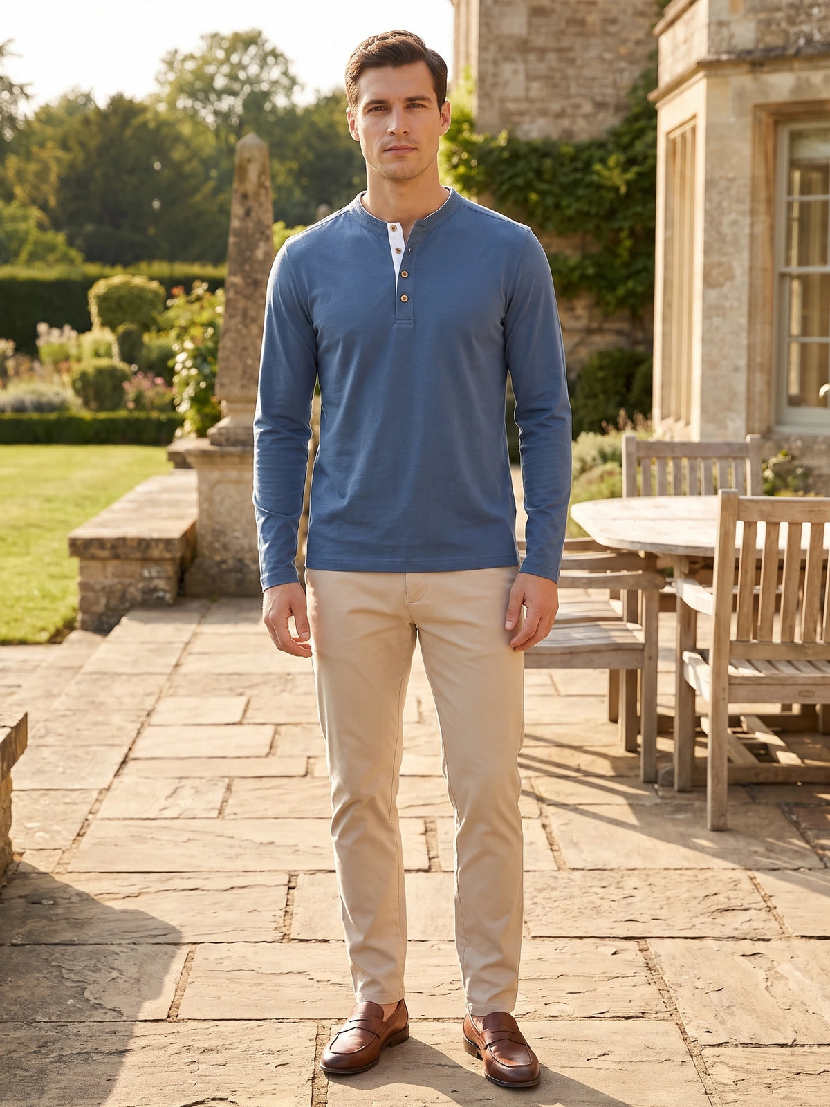 Model wearing a Blue Slim-Fit cotton long-sleeve Henley shirt on a sunlit garden patio beside a stone house.