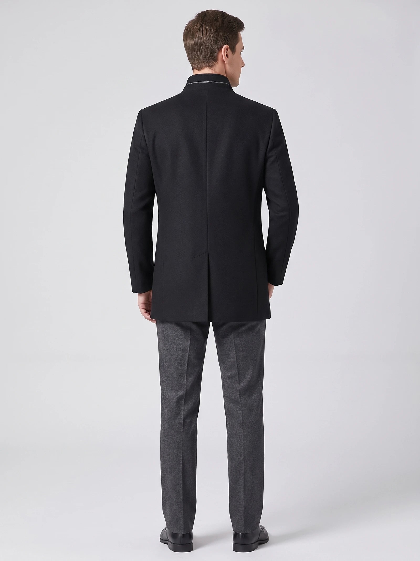 Rear studio view of a model wearing a Black wool overcoat with a stand collar and center back vent.