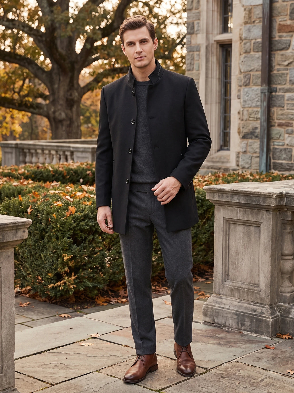Man wearing a Black wool overcoat with a stand collar while walking beside a stone building and autumn trees.