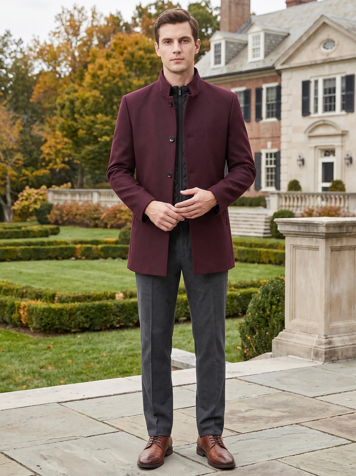 Man wearing a Wine wool overcoat with a stand collar while standing outside a large home.