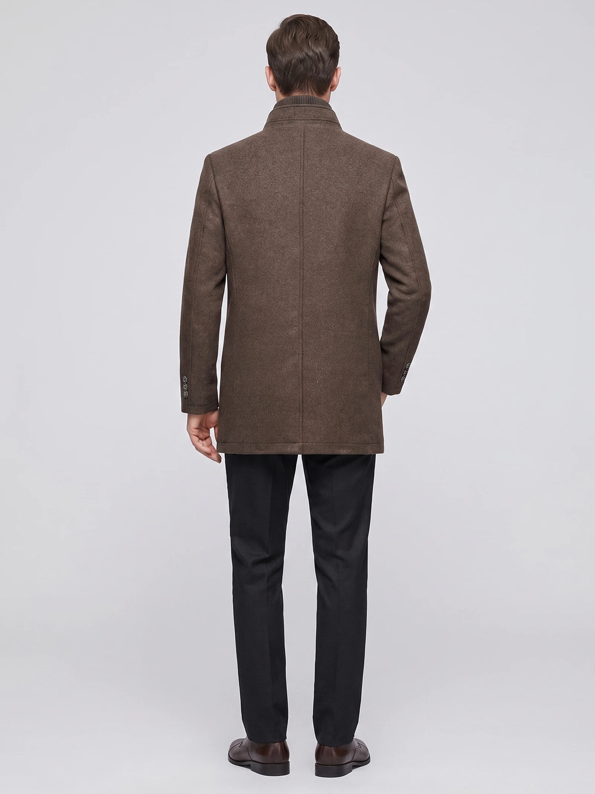 Rear studio view of a model wearing a Brown wool overcoat with a stand collar, back seam, and buttoned cuffs.