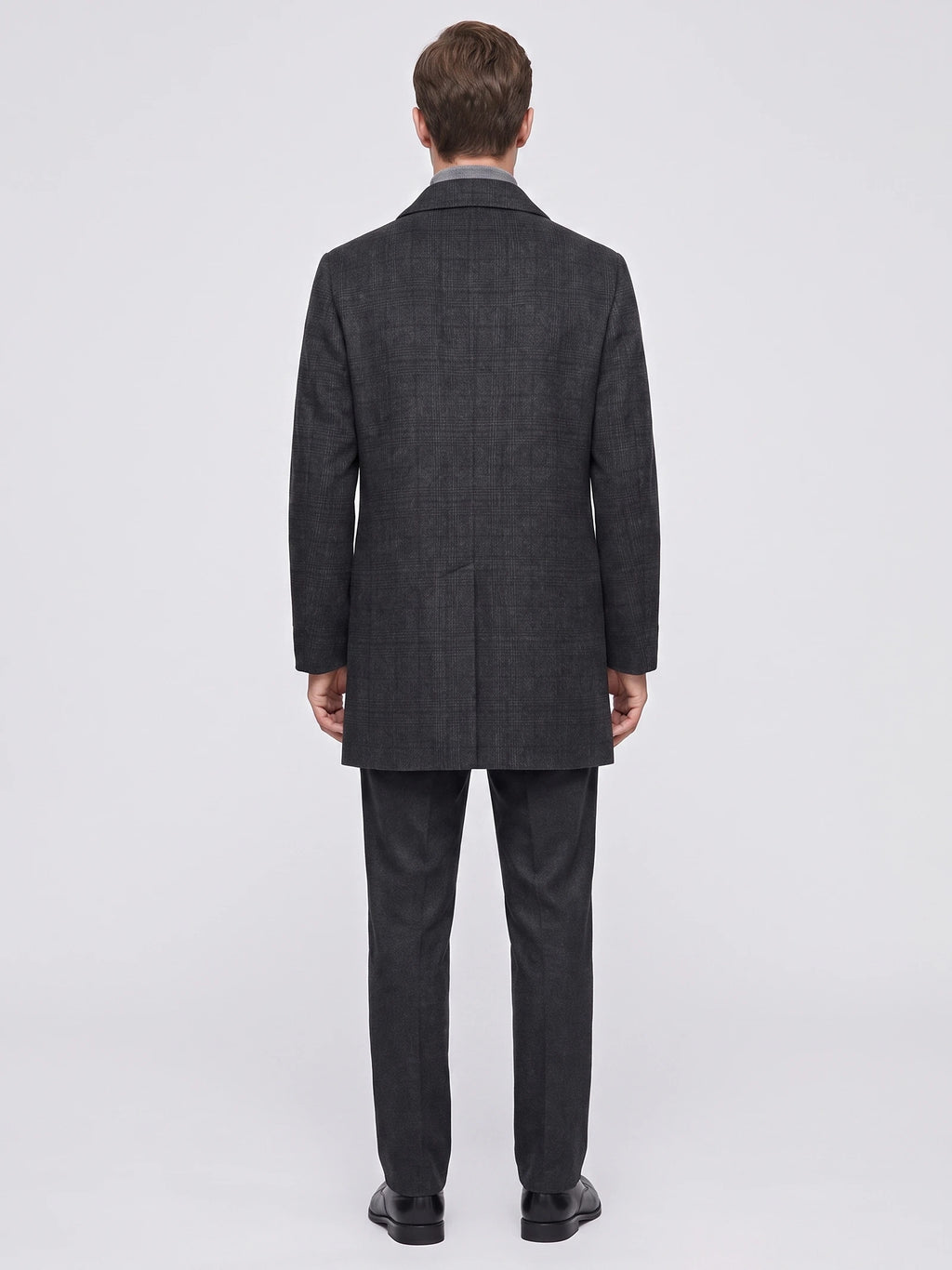 Rear studio view of a model wearing a Black wool trench coat with a subtle plaid texture and back vent.