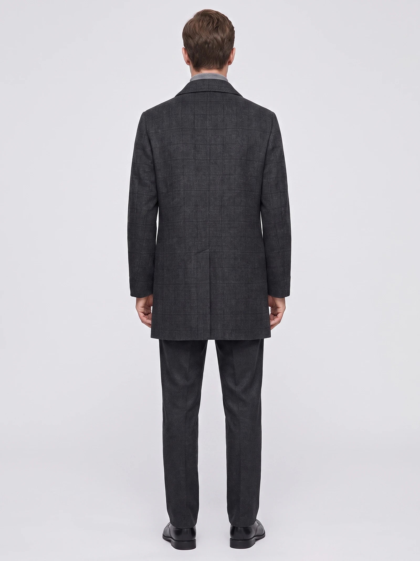 Rear studio view of a model wearing a Black wool trench coat with a subtle plaid texture and back vent.