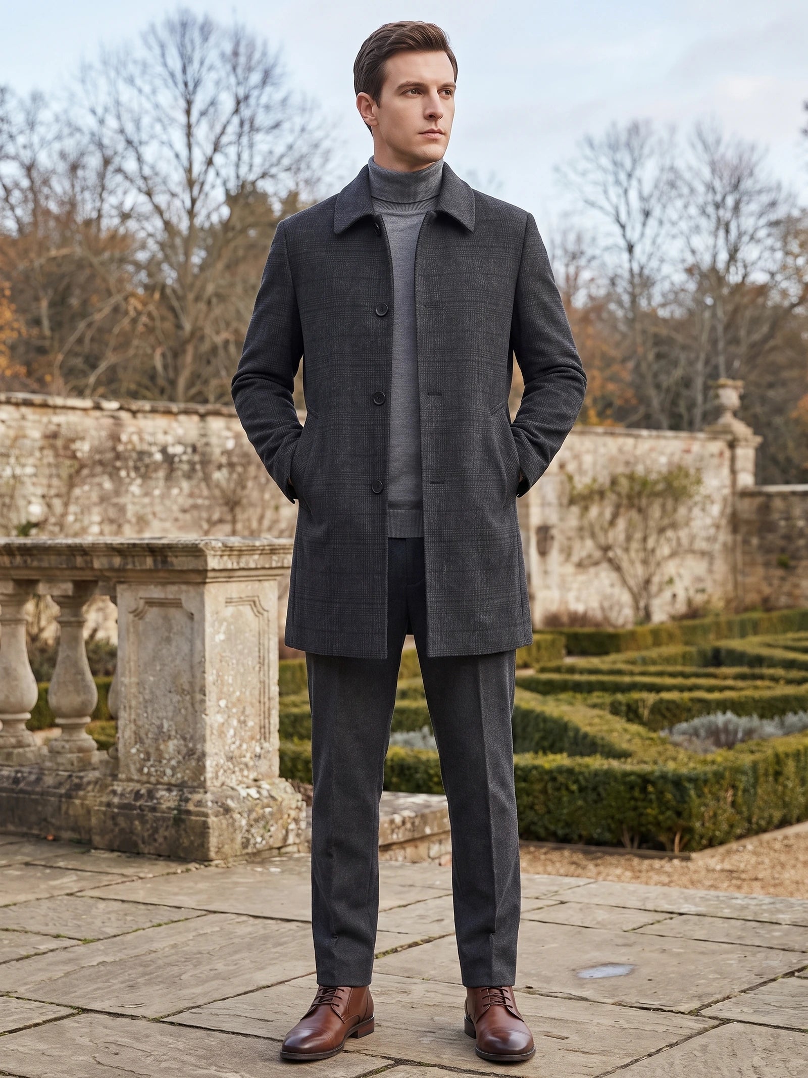 Man wearing a Black Wool Trench Coat with hands in pockets on a stone terrace beside a formal garden.