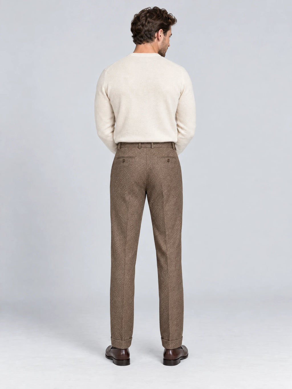 Rear studio view of a model wearing Brown woolen herringbone formal pants with buttoned back welt pockets and cuffed hems.