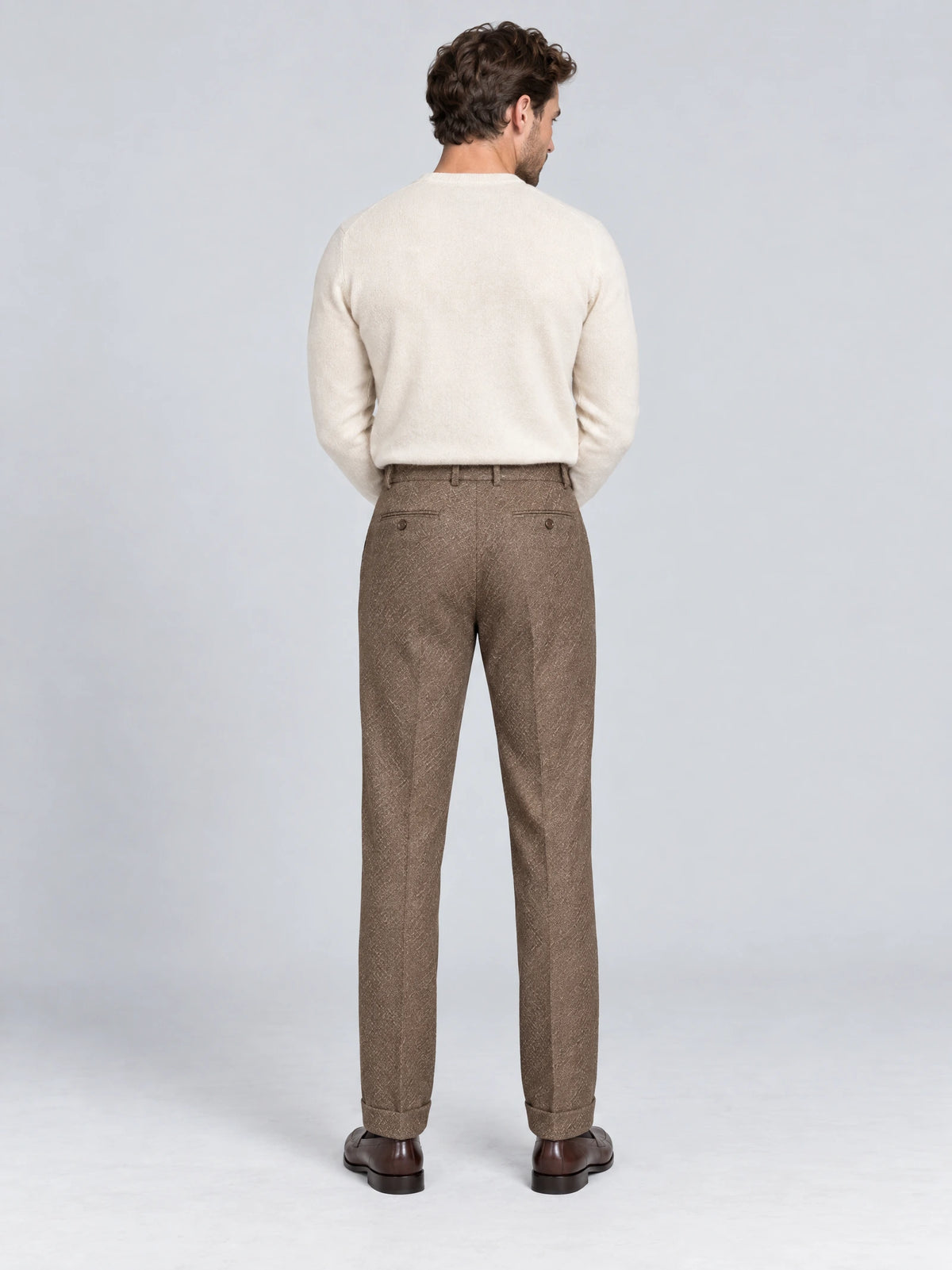 Rear studio view of a model wearing Brown woolen herringbone formal pants with buttoned back welt pockets and cuffed hems.